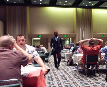 Tom cutting a dashing figure, striding across the board game hall in a suit and Evil Balaclava